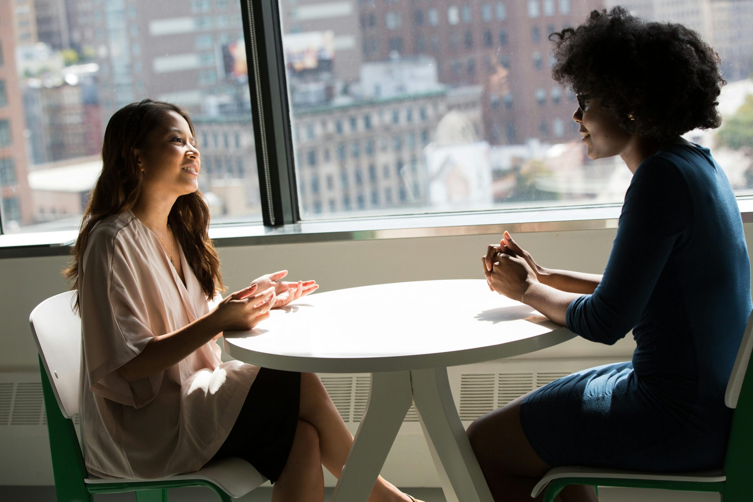 Home Two professional women having a discussion at a desk in a bright office setting.